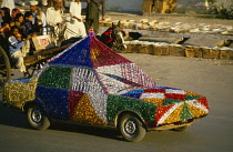 Pakistan, Punjab, Rawalpindi, Wedding car covered in brightly coloured tinsel passing horse drawn cart on street.