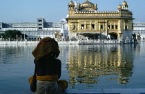 India, Punjab, Amritsar, Pilgrim sitting beside sacred pool looking across to the Golden Temple reflected in rippled water surface.