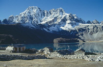 Nepal, Himalayas, Gokyo, Snow covered peaks overlooking lake with stone built houses and livestock enclosures.