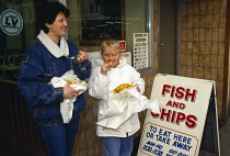 Food, Fast Food, Woman and girl eating fish and chips outside shop with a board advertising opening hours.