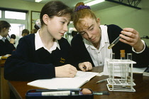 Education, Science Class, Two school girls in uniform at desk examining contents of test tube with open exercise books, pens and pencils in front of them.