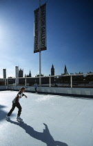 England, London, Boy on an Ice skating rink under a sign for the Saatchi Gallery on the bank of the River Thames with the city skyline in silhouette behind.