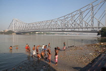 India, West Bengal, Kolkata, Men wash and bathe in the Hooghly River at Malik Ghat in with Howrah Bridge in the background.