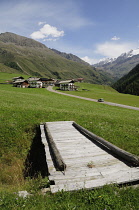 Italy, Trentino Alto Adige, Valle Lunga, wooden bridge, valley views with small village & Otztal Alps in distance.