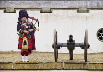 Scotland, Perthshire, Blair Castle, Piper in traditional Scottish outfit playing the bagpipes beside a cannon at the castle.