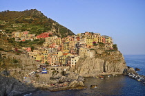Italy, Liguria, Cinque Terre, Manarola, General vista of the town bathed in evening sunshine as seen from Punta Bonfiglio opposite with bathers on rocky ledges.