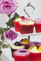 Studio shot of colourfully decorated cupcakes on cake stand with pink roses behind.