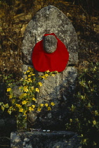 Japan, Religion, Roadside memorial with red cloth tied around stone structure and vase of yellow flowers.