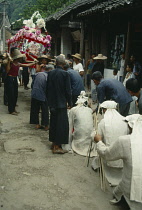 China, Guangxi Province, Fuli, Funeral procession mourners kneeling in front of flower covered coffin carried behind. .