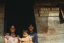 Indonesia, Sumatra, Lake Toba, Samosir Island. Batak Guest House with couple holding child standing in open doorway and welcome sign on the wooden exterior wall.