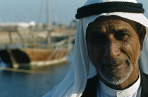 Qatar, Doha, Portrait of a man in the harbour wearing traditional head covering with a dhow at anchor beyond.