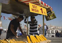 Turkey, Istanbul,  Sultanahmet, Street stall selling freshly grilled corn on the cob outside The New Mosque or Yeni Camii with Galata Bridge behind.