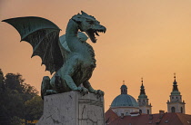 Slovenia, Ljubljana, Dragon Bridge at sunset, One of the dragon statues with the Cathedral of St Nicholas in the background.