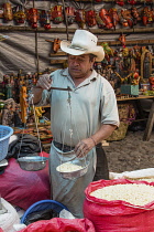 Guatemala, El Quiche Department, Chichicastenango, A Mayan man in modern dress weighs maize or corn kernals for sale with a hand-held balance scale in the Indian market.