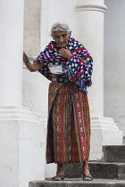 Guatemala, Solola Department, San Pedro la Laguna, An elderly Mayan woman in traditional dress on the steps of the church.  She has her tzute or utility cloth wrapped around her shoulders.