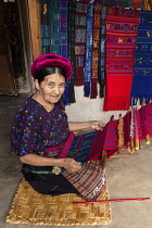 Guatemala, Solola Department, Santa Catarina Palopo, An older Cakchiquel Mayan woman weaves fabric on a backstrap loom while kneeling on the floor of her home.