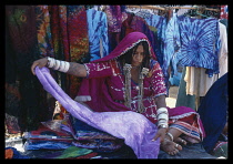 India, Goa, Anjuna, Textile stall at the flea market  female stall holder wearing a pink sari and silver jewellery unrolling a piece of violet coloured cloth.