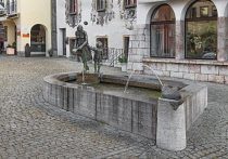 Germany, Bavaria, Berchtesgaden, Mother and Child Fountain also known as the Bucket Fountain with the Monkey Facade of the Deer House in the background.