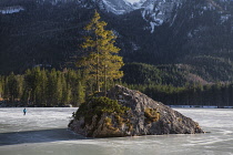 Germany, Bavaria, Berchtesgaden, Berchtesgadener Alps, A tourist ice skating on a frozen section of  Lake Hintersee with a tree perched on a solitary small island and mountains in the background.