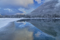 Germany, Bavaria, Berchtesgaden, Berchtesgadener Alps, Partially snow covered and frozen Lake Hintersee with reflection of sky, hillside and clouds in the unfrozen section.