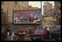 Egypt, Cairo, Cinema hoarding advertising an action film with girl and man selling oranges from a cart near parked cars below.