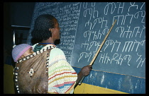 Ethiopia, General, Amharic language teacher indicating script written on blackboard with baby carried in papoose on her back.