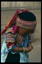 Thailand, North, Children, Young girl from the Lisu tribe wearing brightly coloured traditional dress bending to look at what she holds in her hand.