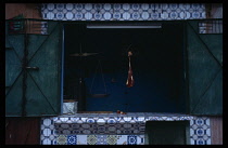 Morocco, Ijoukak, Meat hanging from hook beside set of scales in open hatch of butchers with tiled counter and surround and green painted metal shutters.