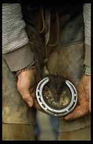 Blacksmith, Farrier, Cropped shot of farrier wearing protective leather apron holding up hoof of horse to show worn metal shoe.