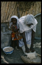 Sudan, East, Food Preparation, Woman grinding dura flour mixed with water to make a flat unleavened bread called kisra. Little girl standing beside her hiding beneath clothing.