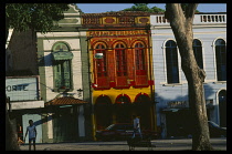 Brazil, Para, Belem, Praca do Relogio.  Late afternoon sunshine on brightly painted building facades with people walking across square and trees in foreground.