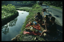 Indonesia, Bali, Mengwi, Family sitting on roadside beside stretch of water with food and flower offerings during festival.