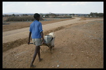 Botswana, Kopong Village, Boy fetching water carried in metal can and plastic container on wheelbarrow.  Dry  dusty unmade road with village huts ahead.