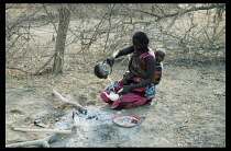 Sudan, South Darfur, General, Baggara Arab woman from the Beni Halba tribe with baby carried in sling on her back  making tea from kettle boiled over open fire.