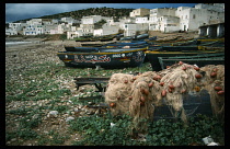 Morocco, Taghzout, Painted fishing boats pulled up onto rocky shore with white painted  flat rooved houses beyond.  Tangled nets heaped in foreground.