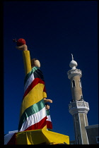 Kuwait, Kuwait City, Statue of mother & child draped in kuwaiti flags during liberation day with Mosque minaret behind.