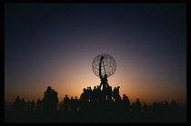 Norway, North Cape, People standing by the globe sculpture looking out to sea towards the midnight sun.