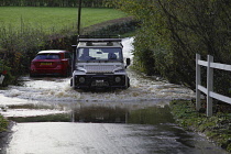 Land Rover Defender 110 passing through water from a swollen stream - tributary of the River Uck - which has flooded the lane between Buxted and Uckfield. Stranded Mercedes car in the background that...