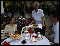 England, Devon, Sidmouth, Couple at outside table laid with traditional cream tea with woman serving cake.  Flowers in window boxes and tubs behind.