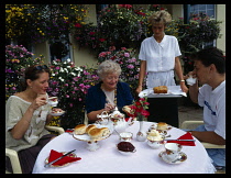 England, Devon, Sidmouth, People at outside table laid with traditional cream tea with woman serving cake.  Flowers in window boxes and tubs behind.