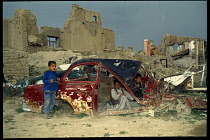 Afghanistan, Kabul, Children playing in bombed out wreckage of car in front of bomb damaged buildings.