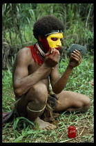 PAPUA NEW GUINEA, Tan, Huli Wigman painting his face in a car wing mirror using modern powder paints.