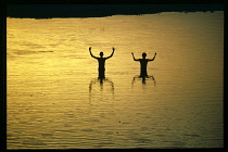 India, Narmada River, Dusk puja or prayers in Holy river.  Two figures standing in river at waist height with raised arms silhouetted against sunset sky reflected in water.