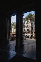 Spain, Balearic Islands, Majorca, Palma de Mallorca, Old Town. View into Placa de La Llotja from inside the medieval guild hall. People at restaurant table under umbrellas in the old market square.