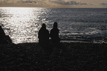 Iceland, Snaefellsnes Peninsula National Park, Djupalonssandur black sand beach. Two people in silhouette sitting on the beach looking out to see.