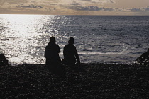 Iceland, Snaefellsnes Peninsula National Park, Djupalonssandur black sand beach. Two people in silhouette sitting on the beach looking out to see.